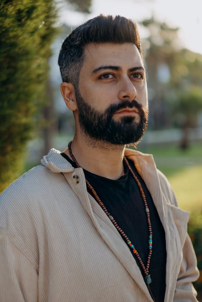 Portrait of a pensive man with beard wearing beads and a beige jacket in natural light.